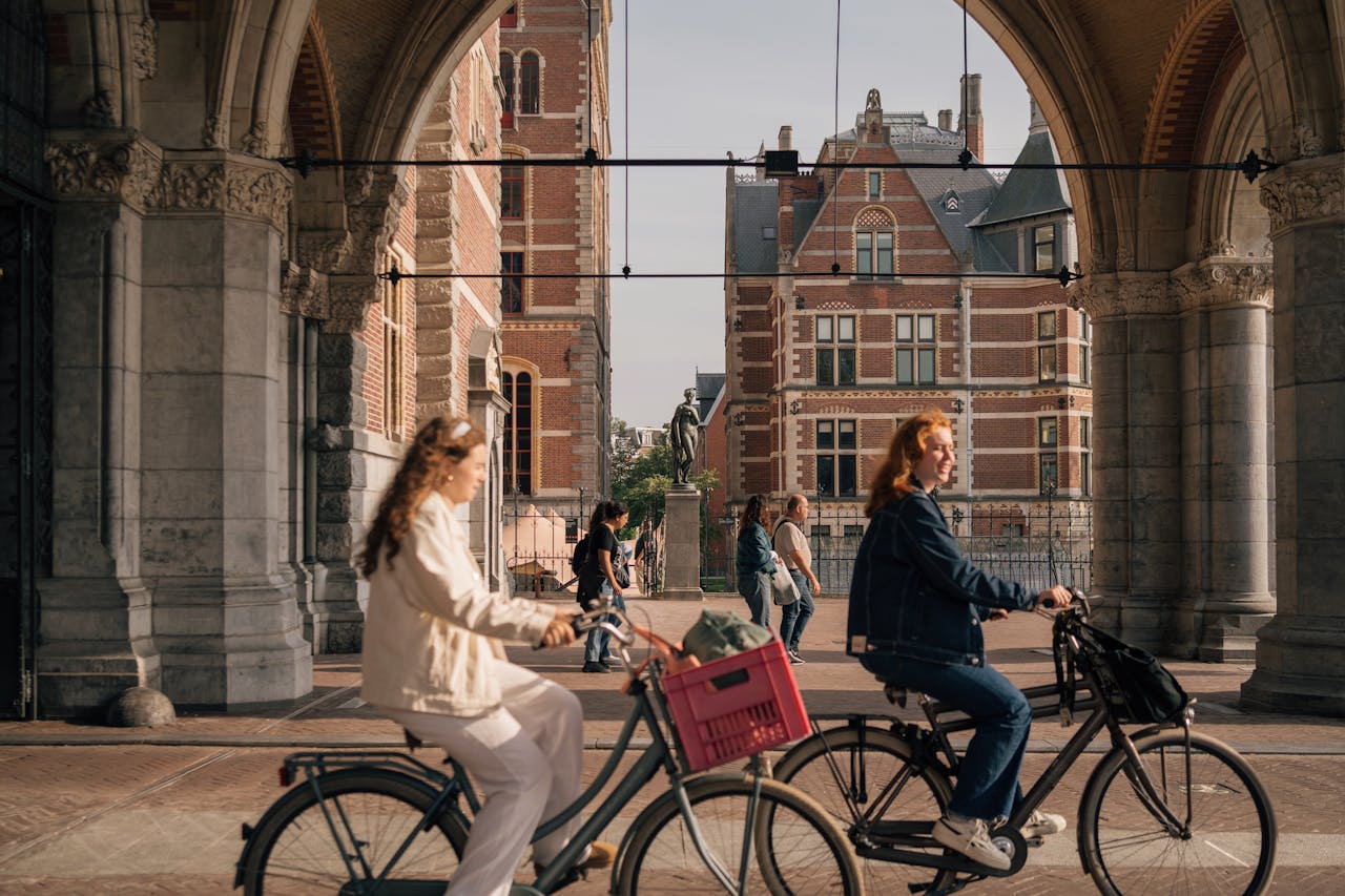Women cycling under an Amsterdam archway with historical buildings in the background.